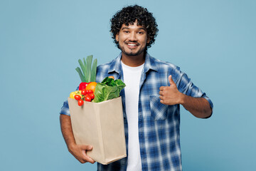 Young smiling happy Indian man wear shirt hold brown craft bag for takeaway mock up with food products show thumb up gesture isolated on plain blue background Delivery service from shop or restaurant