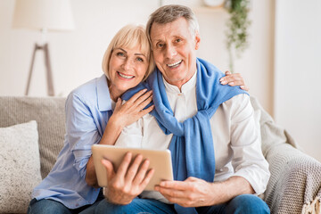 Excited mature couple with digital tablet smiling to camera, woman embracing husband