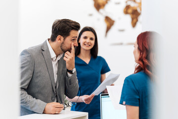 Handsome male patient standing at hospital reception counter and talking with female medical workers. Modern clinic for diagnosis and treatment of eye and sight diseases.