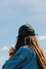 A young woman with long hair, seen from behind, wears a green cap and denim jacket. The soft focus and subtle graininess add an artistic depth to this contemplative scene.
