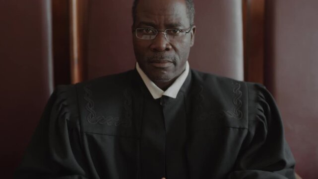 Tilt up portrait of serious impartial African American judge dressed in black robe posing for camera while sitting at desk with open folder in courtroom