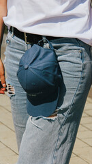 Close-up of a woman holding a blue cap with "located in..." partially visible, paired with ripped blue jeans. The casual and laid-back style emphasizes modern urban fashion trends.