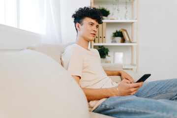 young man using smartphone, relaxed home atmosphere, modern interior, casual clothing, light colors, sitting on couch, thoughtful expression, indoor setting, natural light streaming in, cozy