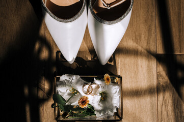 Wedding photography close up of set, accessories, details, top view of beautiful white leather women's shoes of bride, glass box with frame with rings and flowers in hotel room in sunlight.