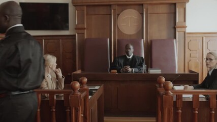 African American policeman leading frustrated Caucasian young male inmate in orange jumpsuit and handcuffs out of courtroom after trial, attorney, prosecutor and judge watching