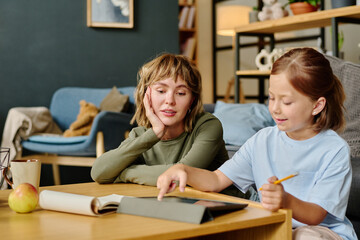 Young Caucasian mom sitting on floor and admiring her daughters art skills, little girl drawing via touchpad