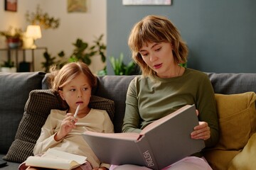 Lovely mom holding huge book with gray cover and reading aloud while her little girl listening to fairytale