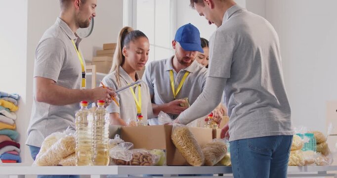 Team of volunteers packing food grocery in donations box for charity food drive. Young cheerful people volunteering in food bank. Humanitarian aid and social help for poor concept. 4k video.