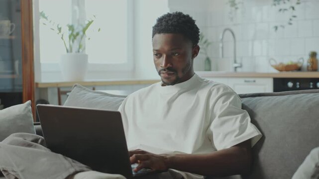 Young focused Black man sitting on couch and typing on laptop, working remotely or surfing the Internet at home