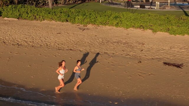 An aerial view shows two girls joyfully running along Oahus sandy shores, as a vibrant sunset illuminates the horizon, reflecting the natural beauty and happiness of their adventure