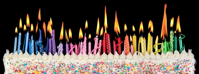 Birthday cake decorated with colorful rainbow sprinkles and white icing topped with lit and burning colorful cake candles in a variety of shapes, sizes and colors.