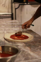 Mano poniendo salsa de jitomate sobre una pizza con fondo rosa claro difuminado. 