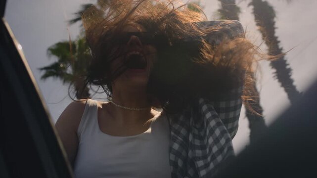 Excited woman enjoys the palm trees of seaside road. A girl leans out of window of rental car on a coastal highway on a sunny day. Hair in the wind. Tropical vacation on the beach of exotic islands