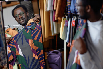 Reflection in mirror of smiling African American man trying vintage summer shirt while shopping at second hand store, copy space