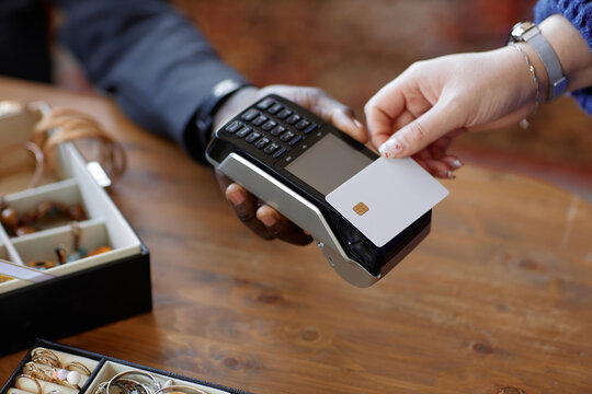 Close up on hand of unrecognizable female customer paying cashless with white mockup credit card and cashier holding POS terminal against wooden checkout counter at second hand shop, copy space