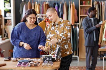 Medium shot of young woman with buzz cut choosing fashion accessories with Middle Eastern female friend dressed in vintage clothes at second hand shop, copy space