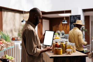 African American man checking the eco friendly products, jars, and groceries on shelves while holding a tablet with a chromakey template. Blank screens for mockups. Sustainable local supermarket.