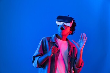 Young African American singer using through VR glasses enjoy singing song with karaoke meta application on blue neon light wall background in virtual reality metaverse fantasy technology. Contrivance.