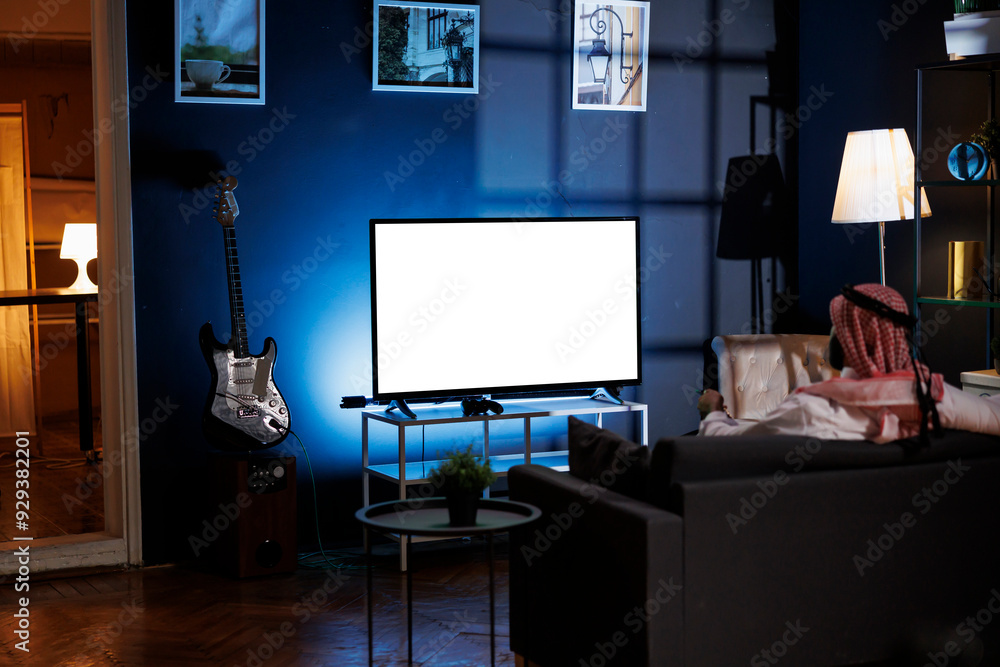 Poster back-view shot of muslim guy in traditional attire seated on sofa in front of television displaying 