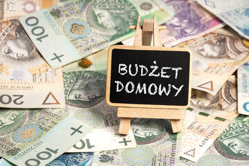 Black writing board on a wooden frame with the inscription "BUDŻET DOMOWY", Polish zloty PLN banknotes scattered in the background (selective focus) translation: HOME BUDGET