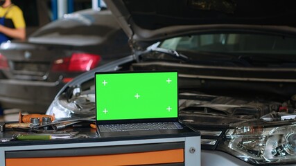 Close up panning shot of green screen laptop in auto repair shop, sitting on work bench next to professional mechanic tools in front of car with hood up waiting to be repaired