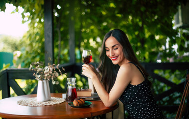 Smiling gorgeous sensual pensive romantic attractive young brunette woman with stylish hairstyle and evening make-up in black dress sitting in cafe and drinking red wine