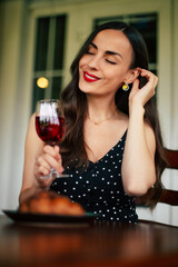 Smiling gorgeous sensual pensive romantic attractive young brunette woman with stylish hairstyle and evening make-up in black dress sitting in cafe and drinking red wine