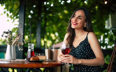 Smiling gorgeous sensual pensive romantic attractive young brunette woman with stylish hairstyle and evening make-up in black dress sitting in cafe and drinking red wine