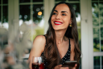 Close up portrait of gorgeous smiling woman in black dress sitting in cafe and using her phone for online shopping, internet searching, chatting and texting with friend while relaxing outside