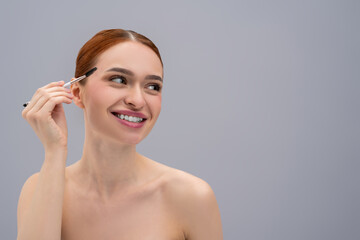 Red-haired woman applying makeup to her face with cosmetic brush isolated over gray background