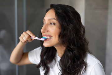 Obraz premium Lady brushing teeth with toothbrush, doing cleaning and whitening toothcare routine, looking at mirror in bathroom. Oral hygiene concept