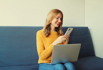 Happy modern young woman working with laptop using phone sitting on a couch in living room at home