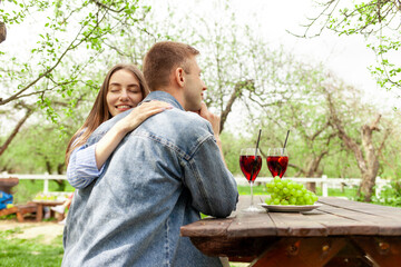 young couple in love hugging and clinking glasses with red wine in the park outdoors, beautiful girl and guy on a date drinking wine and alcoholic cocktails in the garden on a picnic