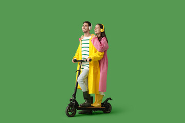 Young couple in raincoats with headphones riding kick scooter on green background