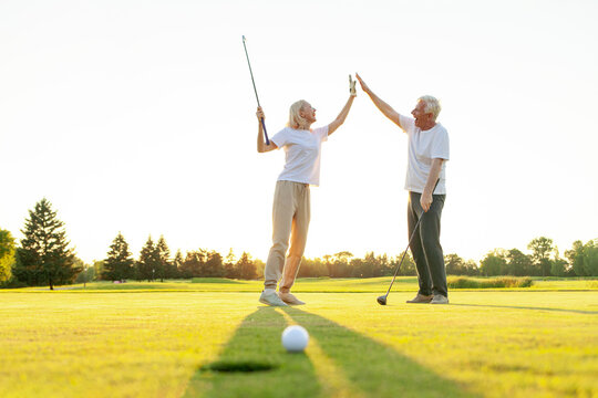 elderly senior couple in uniform celebrating victory and success in golf game and giving high five, old man and woman playing golf on golf course at sunset and doing outdoor sports
