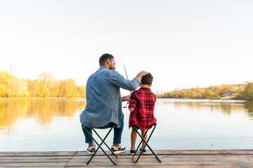 An African American father and his son sit on folding chairs on a wooden pier near a lake. The...
