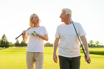 elderly senior couple in uniform holding golf clubs and walking on the background of golf course at sunset and talking, old man and woman walking and talking on golf game and looking at copy space