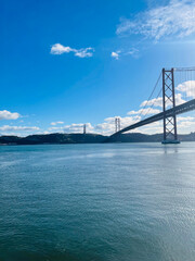 A magnificent bridge stretches across the calm waters under a bright blue sky, connecting the distant city skyline with the natural beauty of the surrounding hills. The perfect blend of urban and natu