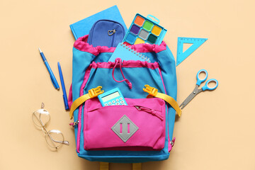 Modern school backpack with different stationery supplies and eyeglasses on beige background