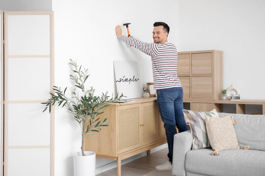 Young man hammering nail into wall for poster at home