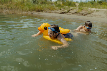 Playful siblings swimming and splashing in the lake on a summer day. The girl is floating with an inflatable yellow duck, while the boy is swimming and teasing his sister