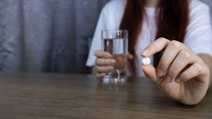 Asian woman's hand holding white pills carefully with fingers and holding glass of water, symbolizing healthcare. Concept of pharmacy, medicine, supplement, vitamins and medical treatment.