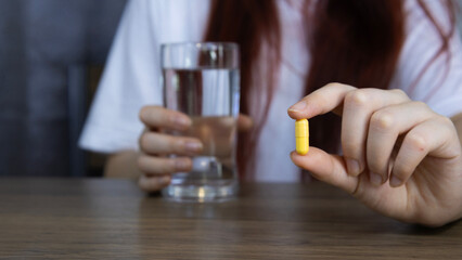 Asian woman's hand holding yellow pill capsule carefully with fingers and holding glass of water, symbolizing healthcare, Concept of pharmacy, medicine, supplement, vitamin and medical treatment.