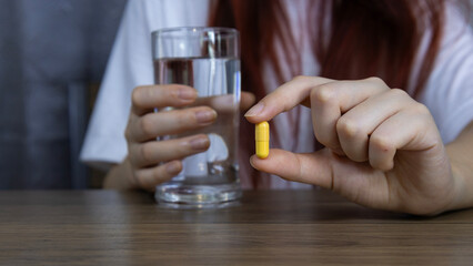 Asian woman's hand holding yellow pill capsule carefully with fingers and holding glass of water, symbolizing healthcare, Concept of pharmacy, medicine, supplement, vitamin and medical treatment.