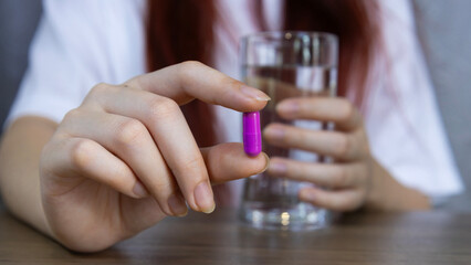Asian woman's hand holding violet pill capsule carefully with fingers and holding glass of water, symbolizing healthcare, Concept of pharmacy, medicine, supplement, vitamin and medical treatment.