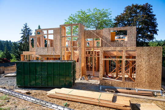 Summer construction season, two story new house build in progress at the framing stage, with large dumpster out front on job site
