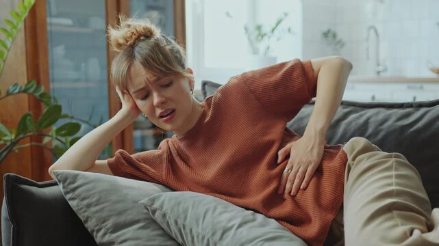 Young woman lying on couch, clutching her abdomen and wincing in pain, feeling stomach ache or cramp during day at home