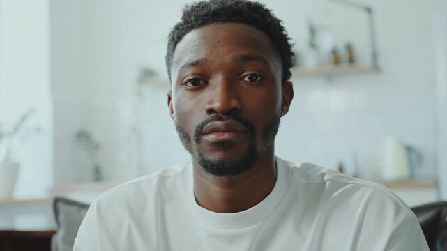 Young African American man with neutral facial expression sitting in the living room and posing at camera. Video portrait