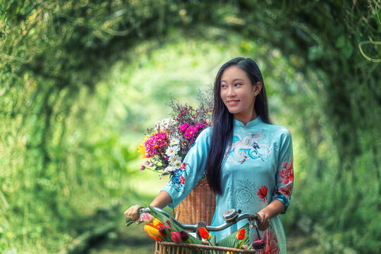 A Moment of Floral Bliss: A young Vietnamese woman in traditional dress smiles serenely next to her bicycle adorned with vibrant blossoms, embodying the beauty and tranquility of rural life. 