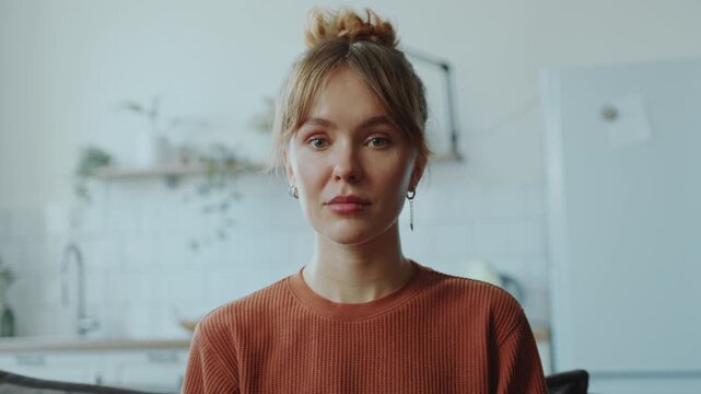 Young beautiful woman with emotionless expression sitting at home and looking at camera. Video portrait